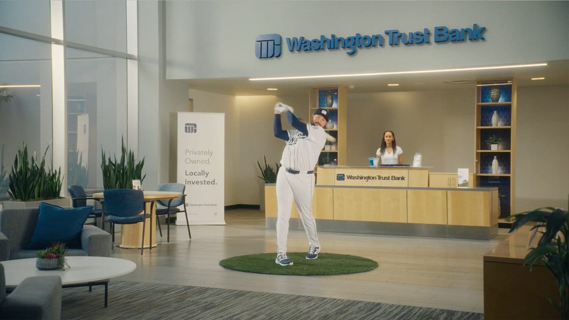 A baseball player taking a full swing on a patch of turf inside an office lobby