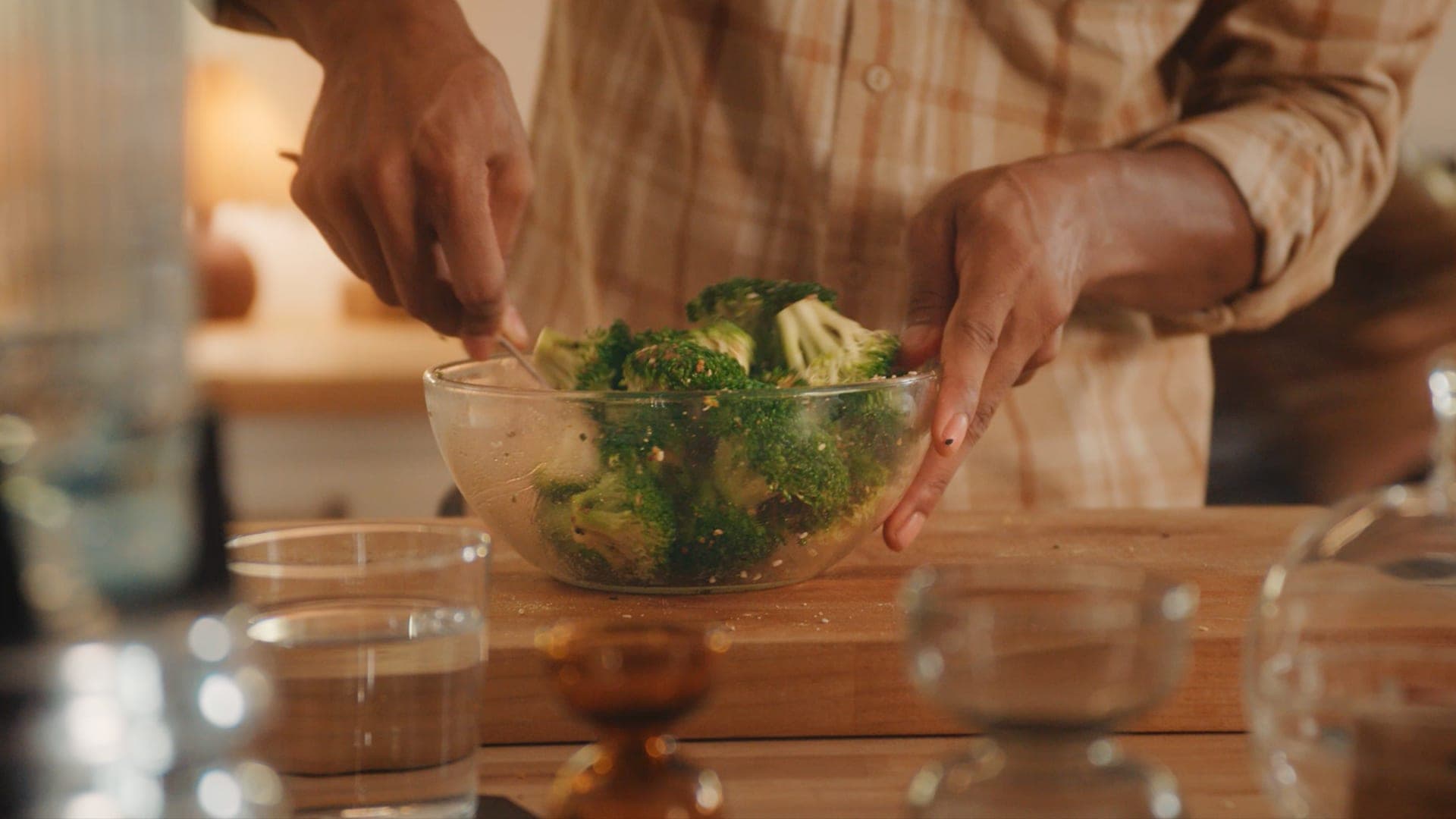 Hands lifting a glass bowl of roasted seasoned vegetables from a wooden counter