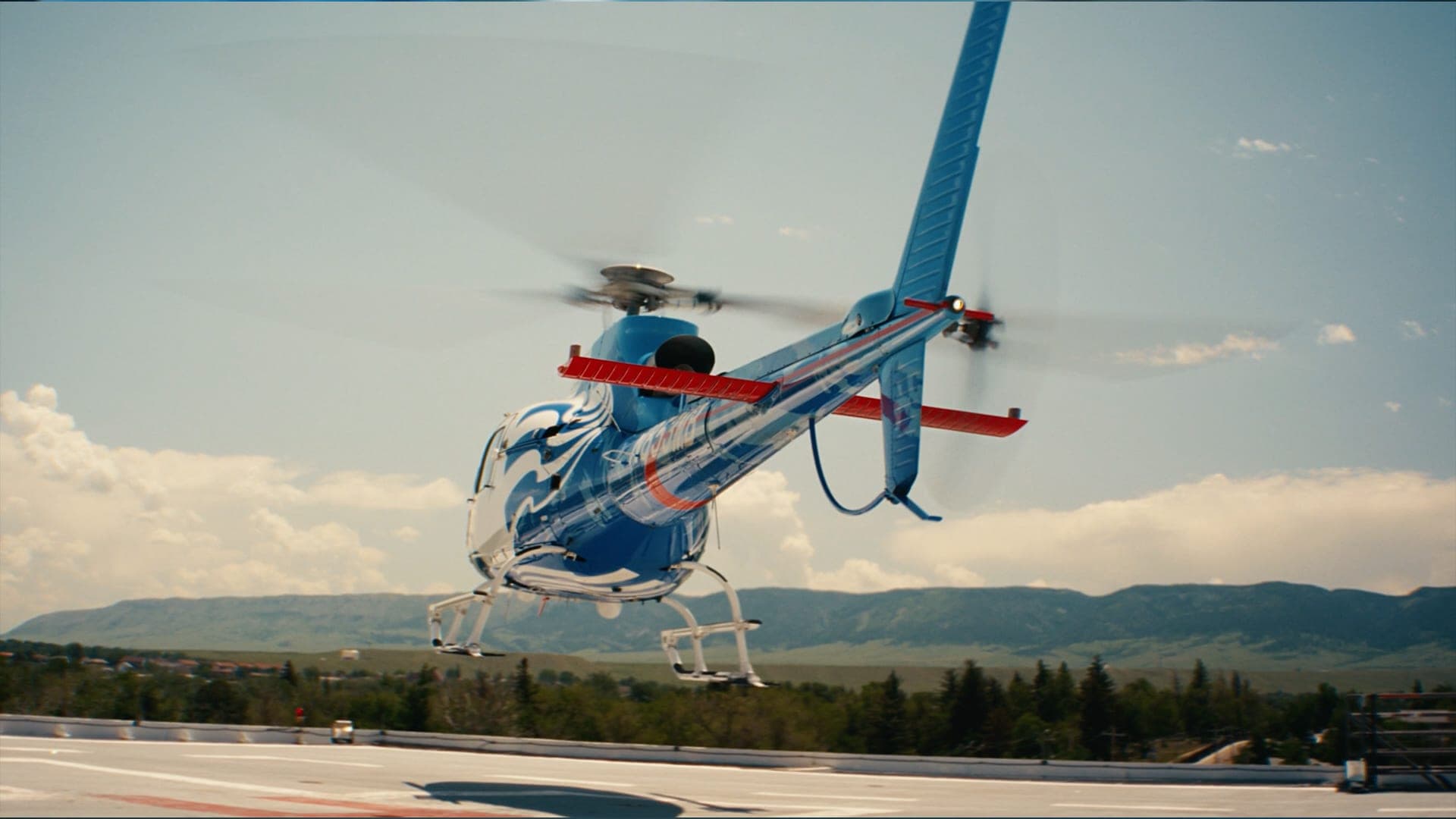 An air-ambulance helicopter lifting off a rooftop helipad, foothills behind it